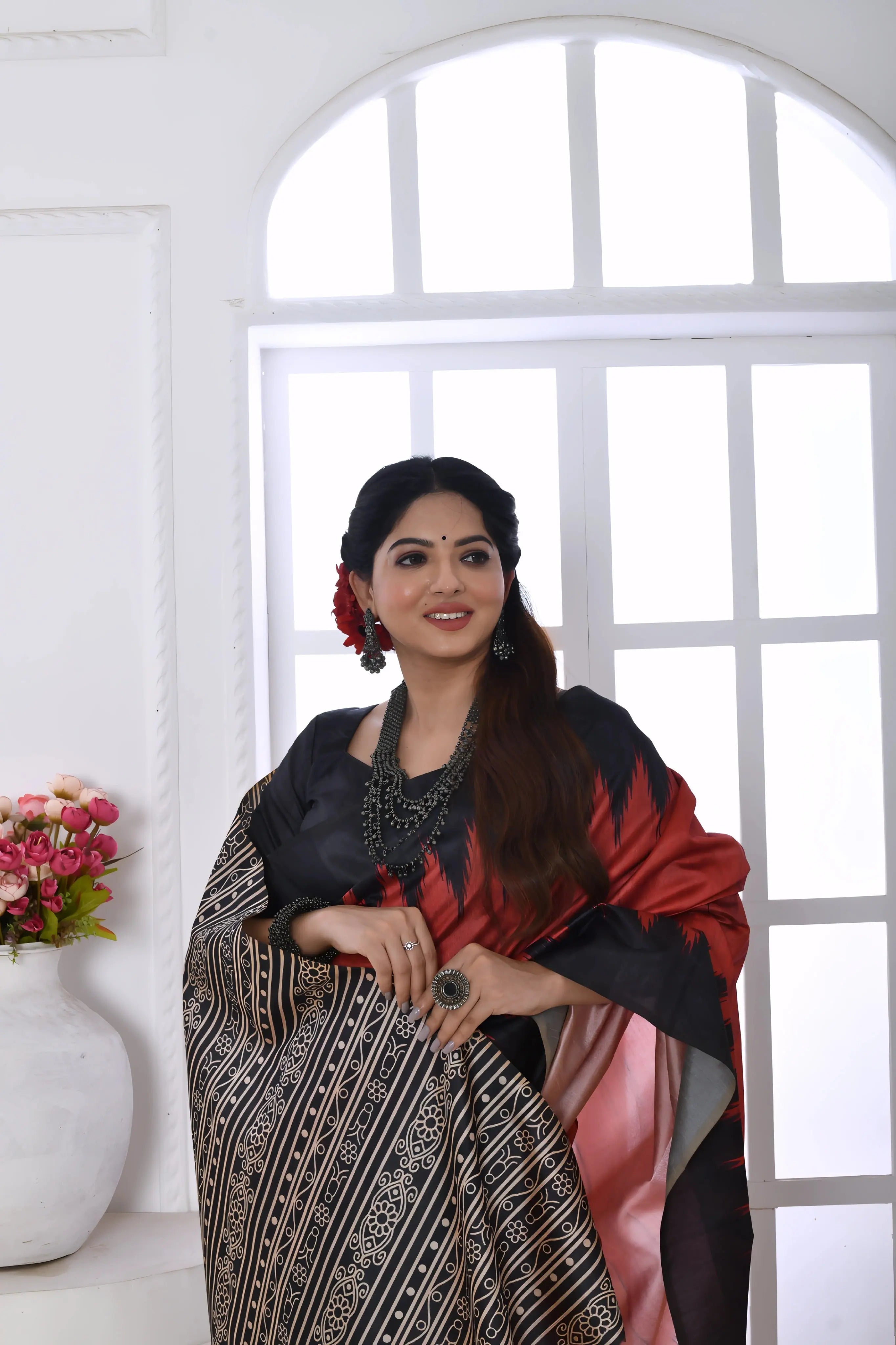 A woman models a striking Terracotta Red Soft Tussar Silk Saree. The saree features a bold black zigzag pattern along the border and a pallu with dense geometric black and white tribal-style print. She wears a black blouse and chunky oxidized silver jewelry, posing indoors by a large window.